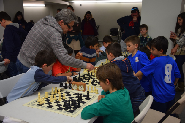 Kinder spielen Schach in einem Wettbewerb, während Erwachsene aus dem Hintergrund und den Seiten zuschauen.