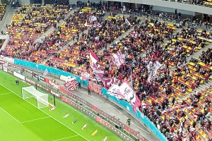Ein großes Stadion voller Zuschauer bei einem Fußballspiel mit einem Tor und Netz auf der linken Seite, einigen Menschen, die vor ihm stehen und sitzen, sowie Tafeln mit Text, Treppen, Geländern und einer Wand im Hintergrund.