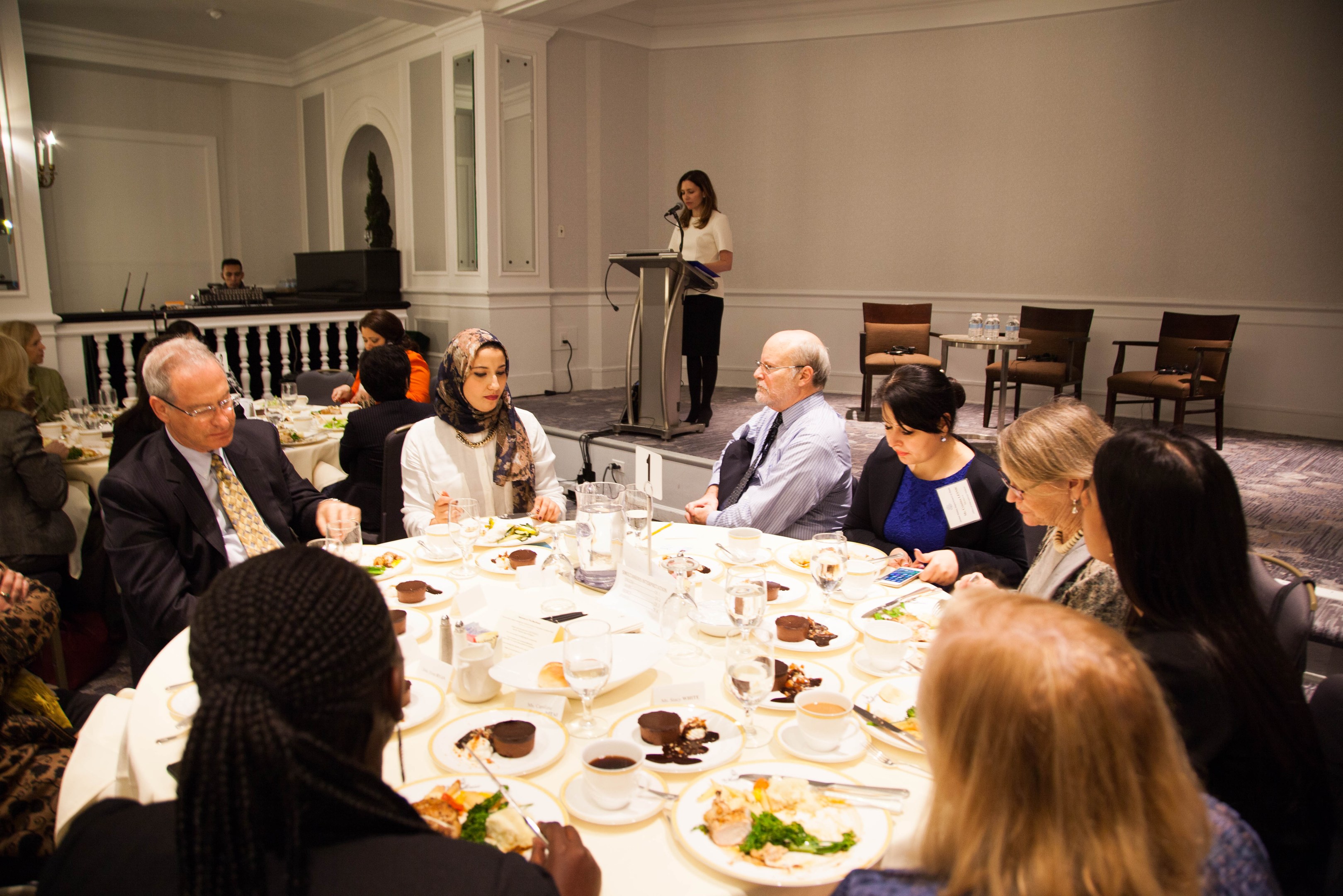 Eine Gruppe von Menschen sitzt um einen Esstisch mit Tellern, Bestecken und Trinkgefäßen, während eine Frau in der Nähe eines Podiums mit einem Mikrofon im Hintergrund zu sein scheint, bei dem es sich um den Women's Health Summit 2018 handeln könnte.