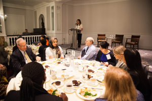 Eine Gruppe von Menschen sitzt um einen Esstisch mit Tellern, Bestecken und Trinkgefäßen, während eine Frau in der Nähe eines Podiums mit einem Mikrofon im Hintergrund zu sein scheint, bei dem es sich um den Women's Health Summit 2018 handeln könnte.