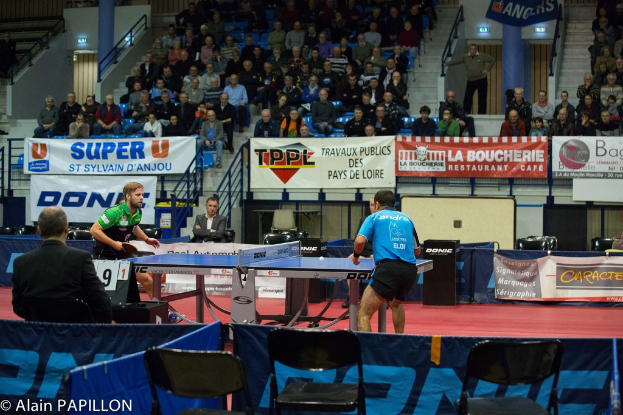 Zwei Männer spielen Tischtennis drinnen mit Zuschauern, die stehen und sitzen, einige leere Stühle und Werbetafeln, mit Text in der unteren linken Ecke.