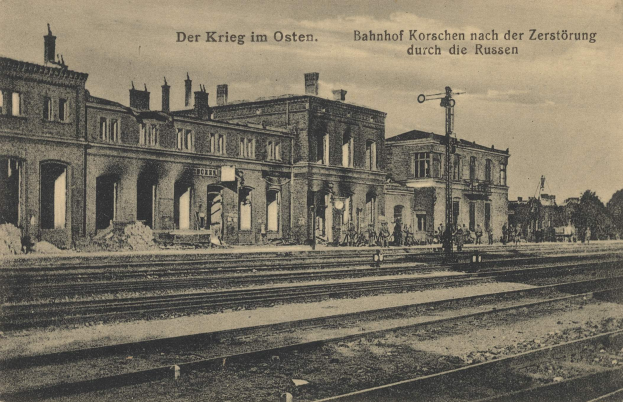 Schwarzes und weißes Foto eines Bahnhofs in Korschen Ost, Deutschland, mit Eisenbahnschienen, Gebäuden, Strommasten, Menschen, Bäumen und Himmel.