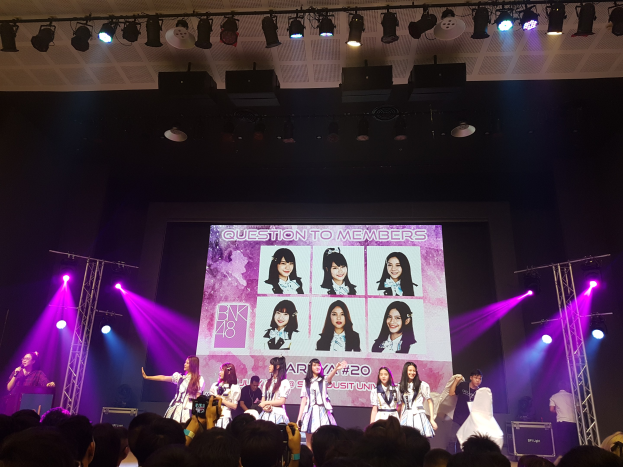 Eine Gruppe von Menschen, die auf einer hell erleuchteten Bühne performen, mit einer Menge darunter, mit einem Menschen mit Mikrofon und einem anderen mit Instrument, und einem großen Bildschirm mit BK48-Bildern im Hintergrund.