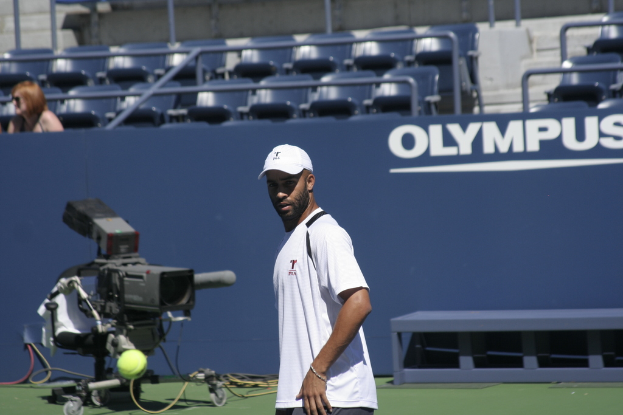 Ein Tennis-Spieler in Aktion mit einem Ball in der Luft, einer Kamera und einem Zaun mit Text dahinter und einer Frau, die im Hintergrund auf der Tribüne sitzt.