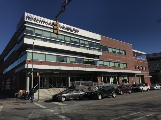 Großes Gebäude, das als New York City Health Care Homeless Center beschriftet ist, mit geparkten Autos, Fußgängern, Straßenmöbeln und Bäumen unter einem klaren Himmel.