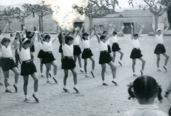 Schwarz-weißes Bild von jungen Mädchen in Schuluniformen, die auf einer von Gebäuden und Bäumen gesäumten Straße tanzen, mit einer Person in Schuhen am unteren Rand.