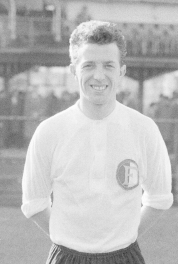 Ein Schwarz-Weiß-Foto eines grinsenden Mannes in T-Shirt und Shorts auf einem Fußballfeld, im Hintergrund ein Gebäude, das mutmaßlich Mario Mandžukić ist.