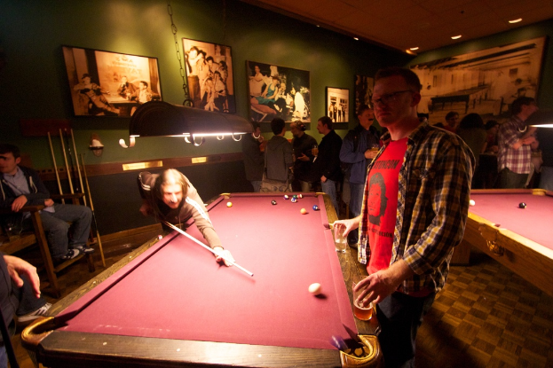 Zwei Personen spielen Snooker an einem von zwei Tischen in einem Club, während mehrere andere im Hintergrund stehen und sich unterhalten, vor einer cremefarbenen Wand mit ein paar Fotorahmen.