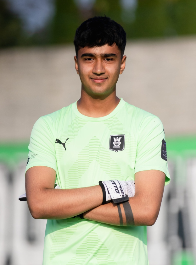 Ein junger Mann in einem grünen Hemd steht auf einem Fußballfeld und verschränkt die Arme, während er lächelt, wahrscheinlich ein Torwart, mit einem unscharfen Hintergrund.