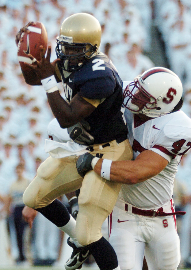 Zwei Fußballspieler in Helmen und Uniformen; einer fängt einen Ball in der Luft, während er von einem anderen getackelt wird.