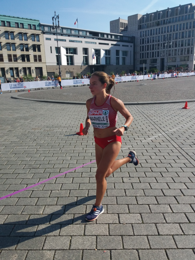 Eine Frau in Sportkleidung beim Laufen eines Marathon, umgeben von Hütchen, einer Schleife, Zuschauern, einem Banner, Straßenlaternen mit Fahnen, Gebäuden und einem bewölkten Himmel.