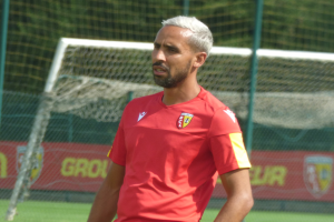 Ein Mann in rotem T-Shirt steht auf einem Fussballfeld und schaut in die Ferne mit einem Netz und einer "Rajasthan FC" Fahne im Hintergrund.