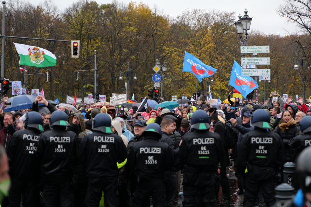 Eine gro├če Gruppe von Polizisten steht vor einer Menge mit Fahnen und Schirmen, mit Laternenmasten, Ampeln, Verkehrszeichen, B├Ąumen und einem klaren Himmel im Hintergrund.