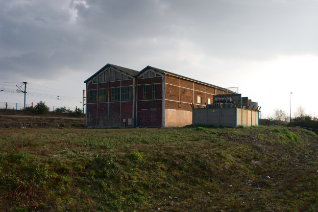 Eine Szene mit Gartenhütten, Transformatoren, Strommasten, Stromkabeln, Zäunen, einer Straßenlaterne, Bäumen und einem bewölkten Himmel.