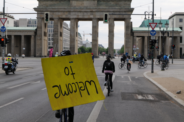 Eine Gruppe von Menschen auf Fahrrädern fährt eine Straße vor dem Brandenburger Tor in Berlin, Deutschland, entlang, trägt Helme und hält ein gelbes Schild hoch, mit Laternenmasten, Verkehrszeichen, Gebäuden, Bäumen und einem klaren blauen Himmel im Hintergrund.