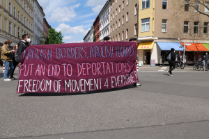 Eine Gruppe von Menschen marschiert mit einem Banner, auf dem "Abolish Borders, Abolish Frontiers, Put an End to Deportations, Freedom of Movement 4 Everyone" steht, durch eine Straße. Im Hintergrund sind Gebäude, Bäume, Fahrräder und eine bewölkte Himmel zu sehen.