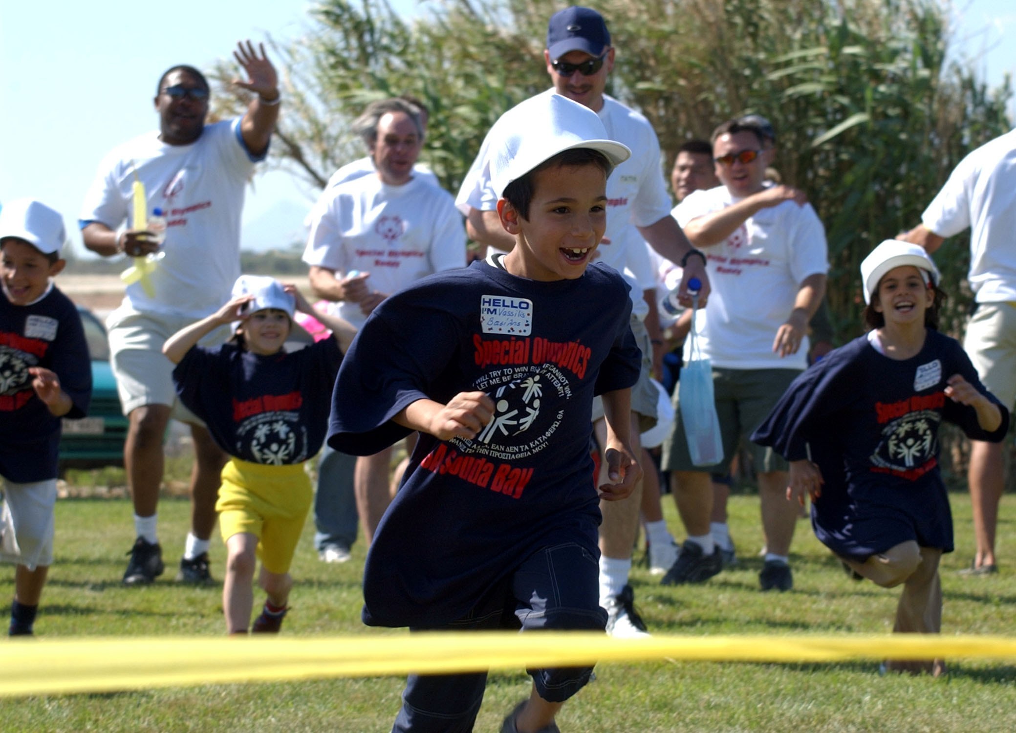 Kinder bei einem Rennen auf einer grünen Wiese mit einem gelben Band im Vordergrund, einige tragen Mützen und Schutzbrillen, andere halten Gegenstände, vor einem Hintergrund aus Bäumen und einem klaren blauen Himmel.