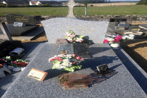 Ein Friedhof mit einem zentralen Grab, geschmückt mit Blumensträußen und einem Buch, umgeben von Häusern, Bäumen und einem klaren blauen Himmel.