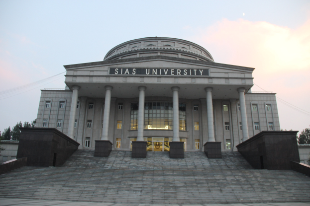 Großes Gebäude mit klassischer Architektur, einschließlich Säulen und Treppen, umgeben von Bäumen unter einem klaren blauen Himmel, mit dem Namen der Universität auf der Fassade.
