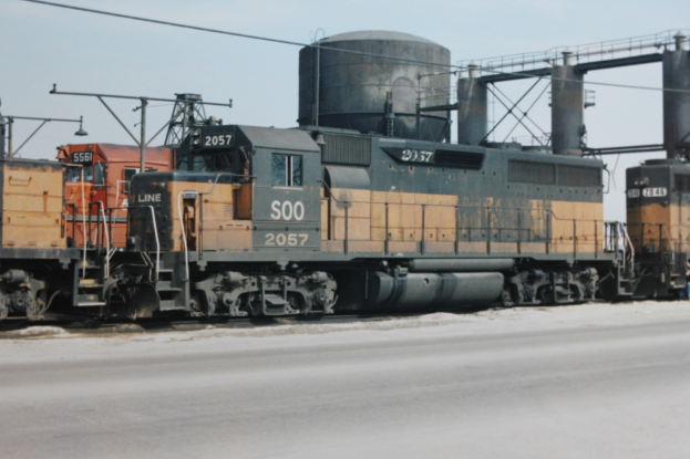Eine Gruppe von Zügen, die parallel auf Schienen fahren, mit einer Straße darunter und einem Himmel im Hintergrund, umgeben von Pfählen, Drähten und anderer Bahninfrastruktur.