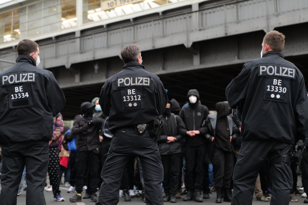 Eine Gruppe uniformierter Polizisten steht vor einer Menge von Menschen in schwarzen Uniformen und Masken, im Hintergrund eine Brücke und ein Gebäude, was auf eine städtische Umgebung hinweist.
