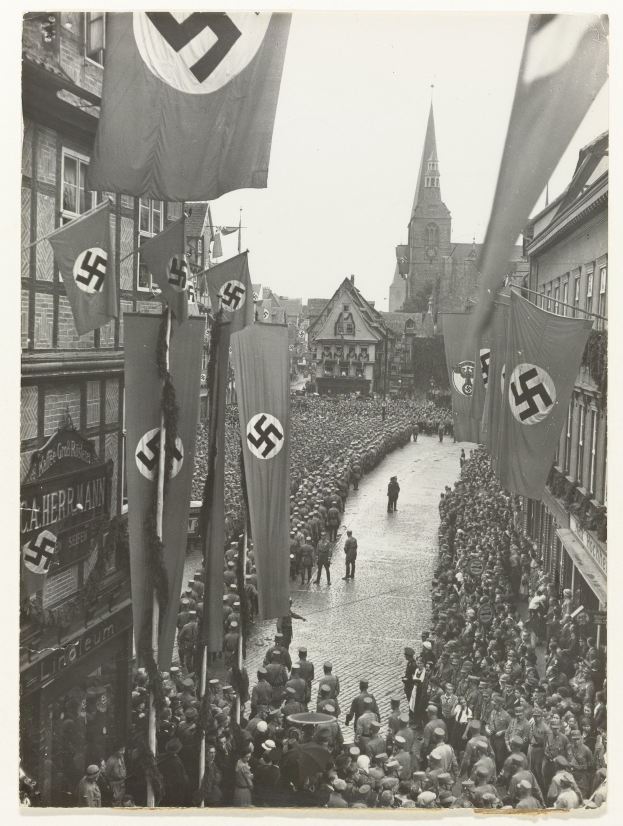 Eine gro├če Menge Menschen, die mit Nazi-Flaggen die Stra├če entlangmarschieren, mit Geb├Ąuden und einem klaren Himmel im Hintergrund, dargestellt in Schwarz-Wei├č.