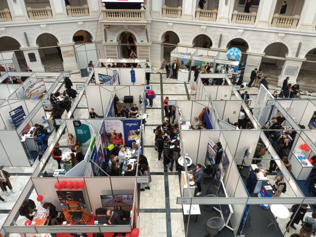 Große Besuchergruppe auf einer internationalen Fachmesse mit Ständen, Bannern, Tischen, Stühlen und Luftballons in einem Raum mit Bogengängen, Säulen und einem Bildschirm im Hintergrund.