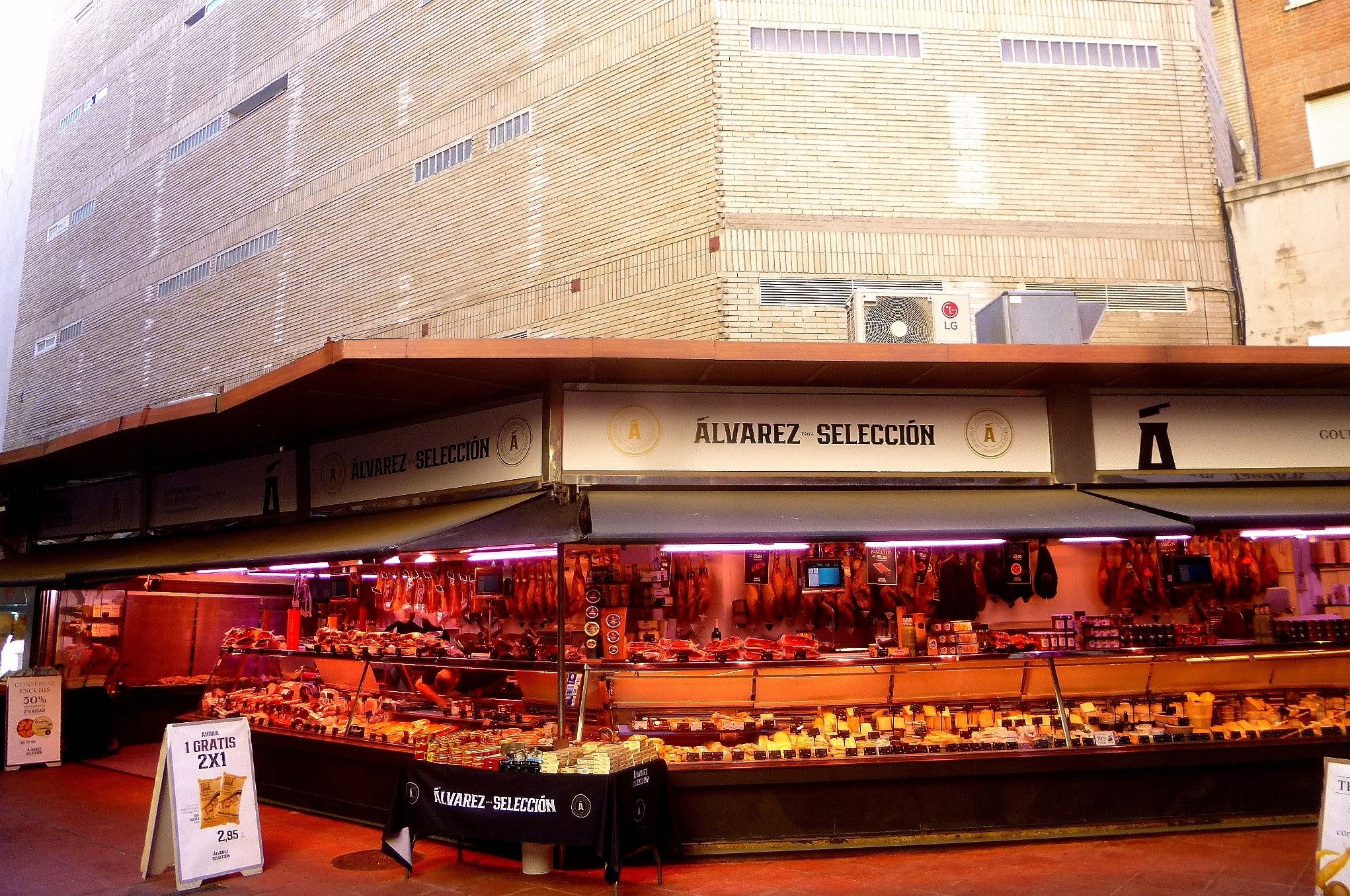 Ein belebter Markt zeigt verschiedene Lebensmittel unter Lichtern, mit Texttafeln und Gebäuden im Hintergrund unter einem klaren blauen Himmel.