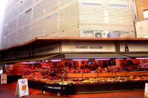 Ein belebter Markt zeigt verschiedene Lebensmittel unter Lichtern, mit Texttafeln und Gebäuden im Hintergrund unter einem klaren blauen Himmel.