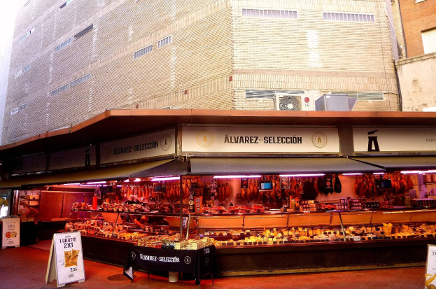 Ein belebter Markt zeigt verschiedene Lebensmittel unter Lichtern, mit Texttafeln und Gebäuden im Hintergrund unter einem klaren blauen Himmel.
