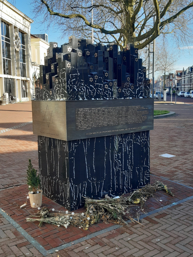 Denkmal in einem Stadtplatz für Holocaust-Opfer mit einem Baum davor, einem Topfgewächsh auf dem Boden, verstreuten trockenen Blättern und Gebäuden, Fahrzeugen, Masten und Bäumen im Hintergrund.