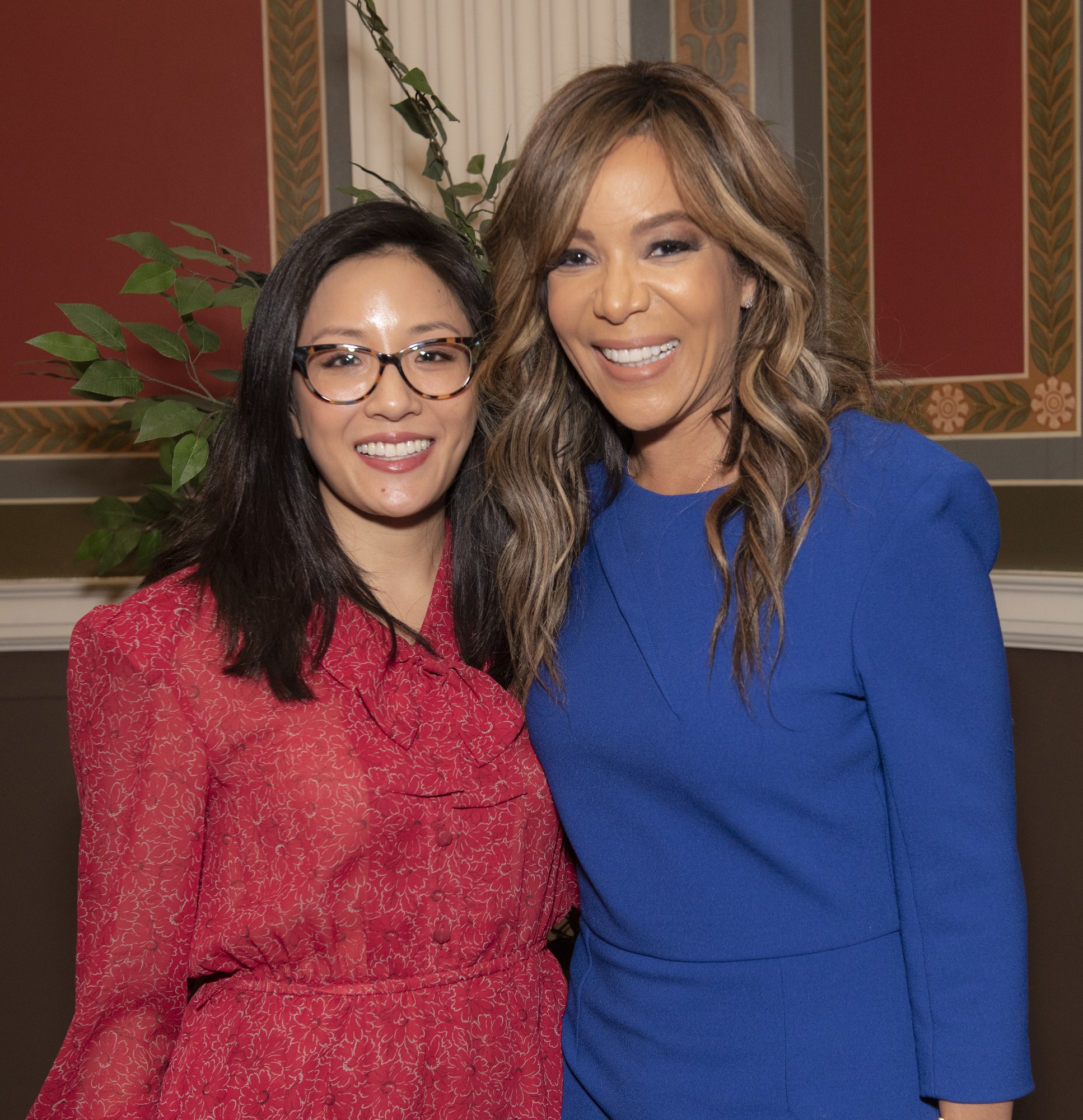 Zwei Frauen, die bei der 2019er Women in Film Pre-Oscar-Luncheon zusammen lächeln, eine in einem blauen Kleid und die andere in einem roten Kleid mit Brille, vor einer Wand mit einer Pflanze und einem Vorhang im Hintergrund.