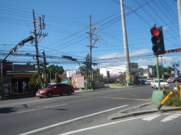 Ein rotes Auto fährt auf einer Straße mit einer Ampel, Strommasten und Kabeln neben Gebäuden, Bäumen und einem klaren blauen Himmel im Hintergrund.