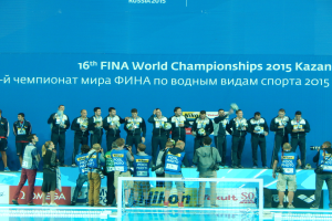 Eine Gruppe von Menschen steht oben auf einem Swimmingpool, einige halten Kameras, mit einem Netz im Vordergrund und einer Flagge mit der Aufschrift "FINA World Championships 2015 Kazan" im Hintergrund.