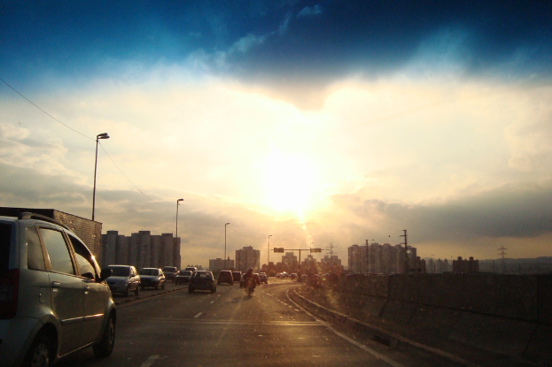 Eine Straße mit wenigen fahrenden Autos, gesäumt von Gebäuden auf beiden Seiten, unter einem Himmel mit der Sonne.