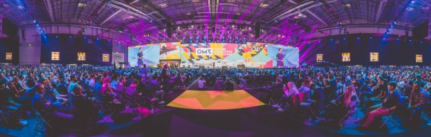 Eine große Menschenmenge, die auf Stühlen sitzt und einer hell erleuchteten Bühne mit mehreren stehenden Personen zugewandt ist, wahrscheinlich auf einem Online-Marketing-Rockstars-Event, umgeben von Bildschirmen.
