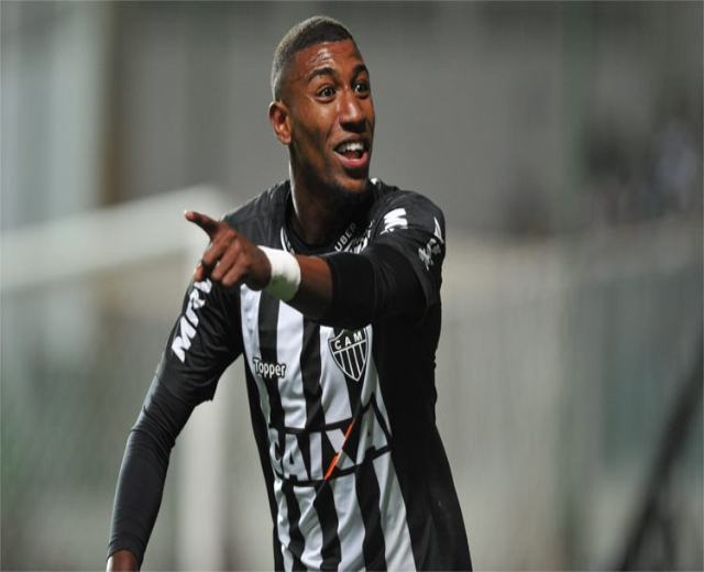 Fußballspieler in einem schwarzen und weißen gestreiften Trikot, der grinst und ein Tor feiert, mit einem unscharfen Hintergrund.