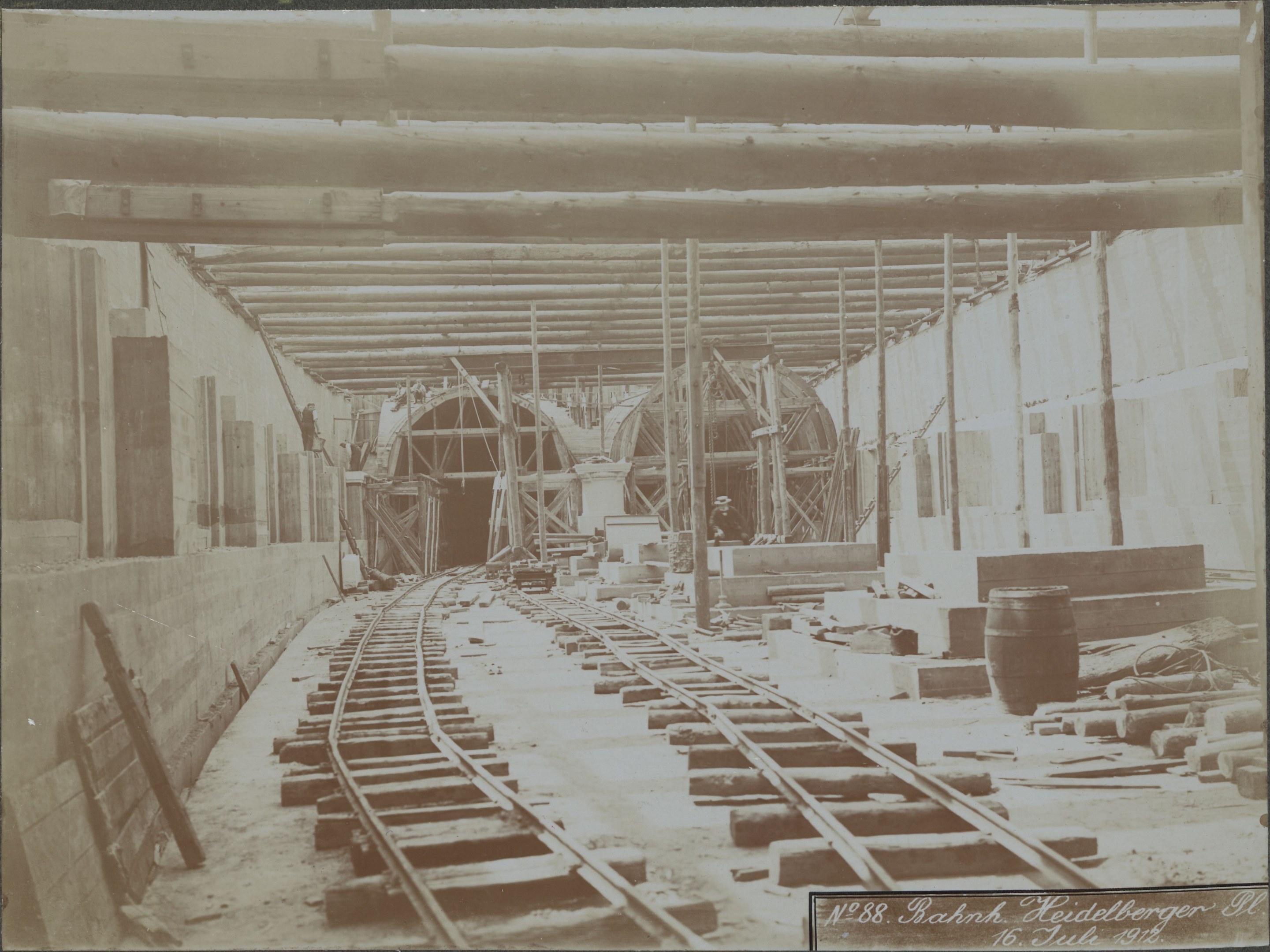 Altes Schwarz-Weiß-Foto einer Bahngleise in einem Gebäude unter Bau, mit verstreuten Holzgegenständen, Fässern, Wänden, Pfählen und anderem Gerümpel und Text unten.