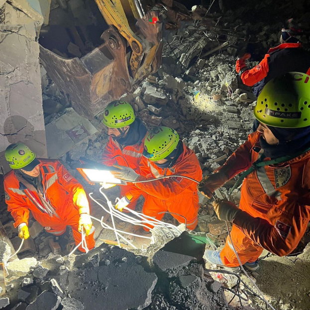Eine Gruppe von Menschen in orangefarbigen Uniformen und grünen Helmen bei der Arbeit an Schutt von einem eingestürzten Gebäude, mit einem Kran im Hintergrund.