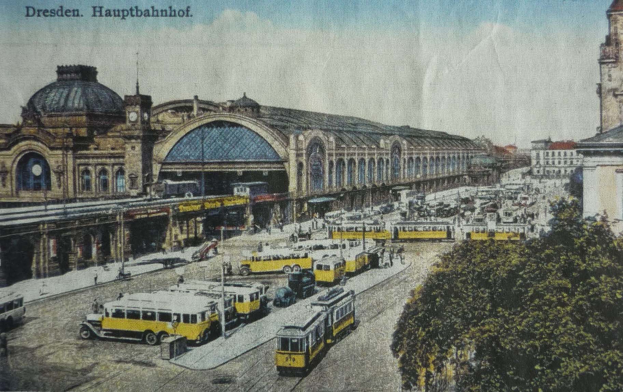 Eine alte Postkarte, die einen belebten Dresdner Bahnhof mit Fahrzeugen, Menschen, B├Ąumen, Geb├Ąuden und einem bew├Âlktem Himmel zeigt, mit der Aufschrift "Dresden Hauptbahnhof" ├╝ber dem Bild.