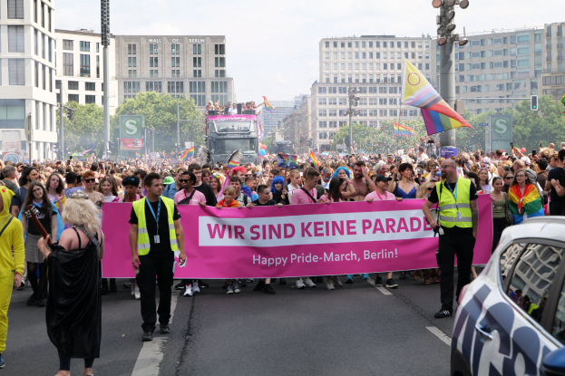 Eine große Gruppe von Menschen nimmt an der 'Happy Pride March Berlin'-Parade teil, hält ein Banner und geht eine Straße mit einem geparkten Auto auf der rechten Seite, Gebäuden und Bäumen im Hintergrund unter einem klaren blauen Himmel entlang.