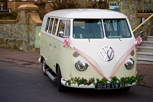 Ein weißer und rosafarbener VW-Bus mit floralen Verzierungen, der vor einer Steinmauer mit Treppe und einem Gebäude im Hintergrund geparkt ist.