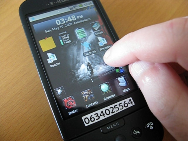 Eine Person nutzt ein Mobiltelefon auf einem Tisch, mit mehreren Apps auf dem Bildschirm und einem Zettel mit einer Nummer daran.