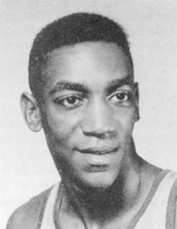 Ein Schwarz-Weiß-Foto eines grinsenden Mannes in einem weißen "Basketball Hall of Fame"-Trikot mit zurückgekämmtem Haar und einem selbstbewussten Gesichtsausdruck.
