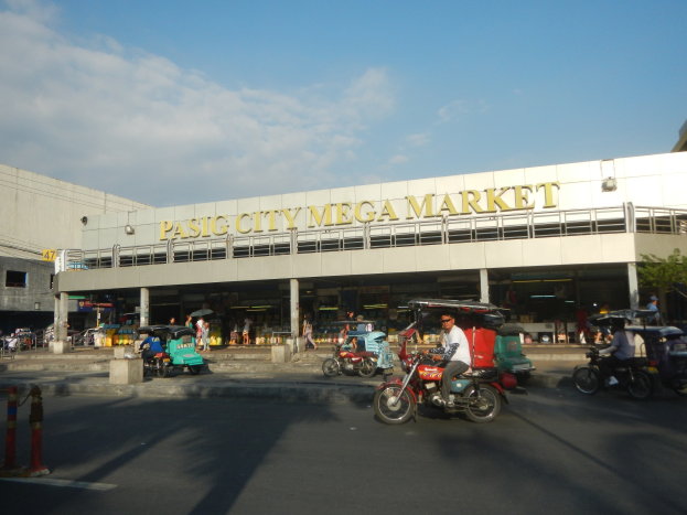 Ein belebter Pasig City Mega Market mit Fahrzeugen auf der Straße, Menschen, die gehen, einem Gebäude mit Text im Hintergrund und einer bewölkten Himmel.