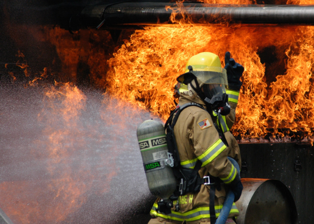 Feuerwehrmann in Schutzausrüstung sprüht Wasser auf ein brennendes Rohr mit einem Schlauch.