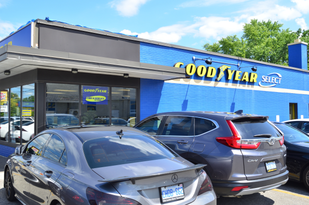Ein Goodyear Select Autohaus mit mehreren geparkten Autos, das ein Gebäude mit Text und Bäume mit einem bewölkten Himmel im Hintergrund zeigt.