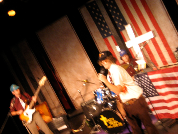 Eine Gruppe von Menschen, die Musikinstrumente spielen und von zusätzlicher Ausrüstung umgeben sind, mit einer amerikanischen Flagge im Hintergrund und einer Wand im Hintergrund.