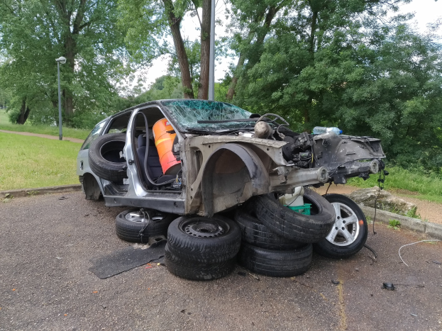 Ein umgestürzter Wagen am Straßenrand umgeben von Trümmern, Gras, Pflanzen, Bäumen, einer Straßenlaterne und einem bewölkten Himmel.