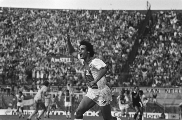 Ein Schwarz-Weiß-Foto eines Fußballspielers, der nach einem Torjubel auf dem Boden steht und von einer Gruppe Menschen umgeben ist, im Hintergrund ein Zaun, Tribünen und eine große Menschenmenge.